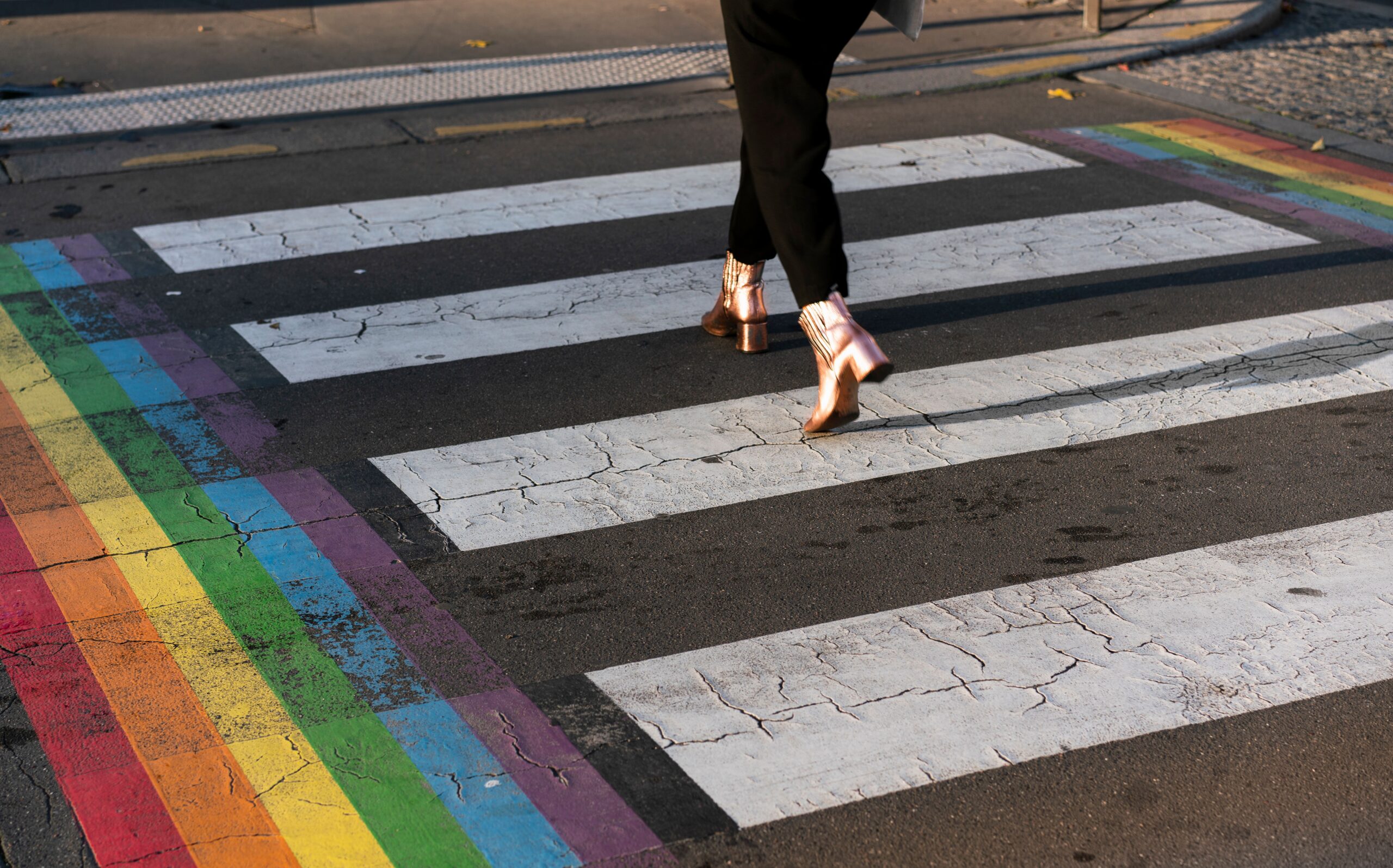 Someone in copper boots walking across a city crosswalk edged with a rainbow stripe